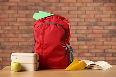 Photo of Backpack, paper planes, book, lunchbox and apple on wooden table against red brick wall