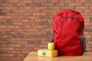 Photo of Backpack, lunchbox and apple on wooden table against red brick wall. Space for text