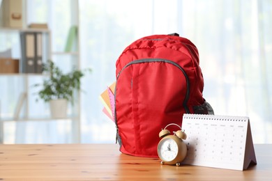Photo of Red backpack with different stationery, calendar and alarm clock on desk in classroom