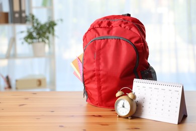 Photo of Red backpack with different stationery, calendar and alarm clock on desk in classroom