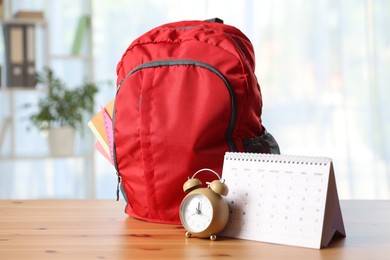 Photo of Red backpack with different stationery, calendar and alarm clock on desk in classroom