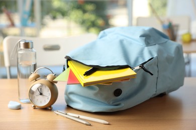 Photo of Backpack with different stationery, alarm clock and bottle of water on desk in classroom, closeup