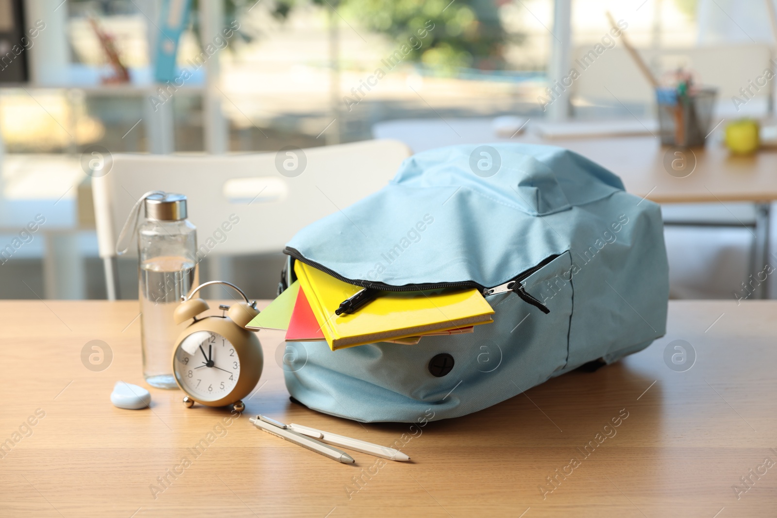 Photo of Backpack with different stationery, alarm clock and bottle of water on desk in classroom, closeup