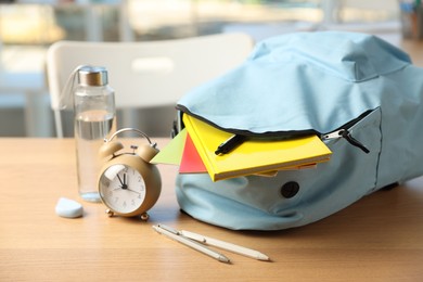 Photo of Backpack with different stationery, alarm clock and bottle of water on desk in classroom, closeup