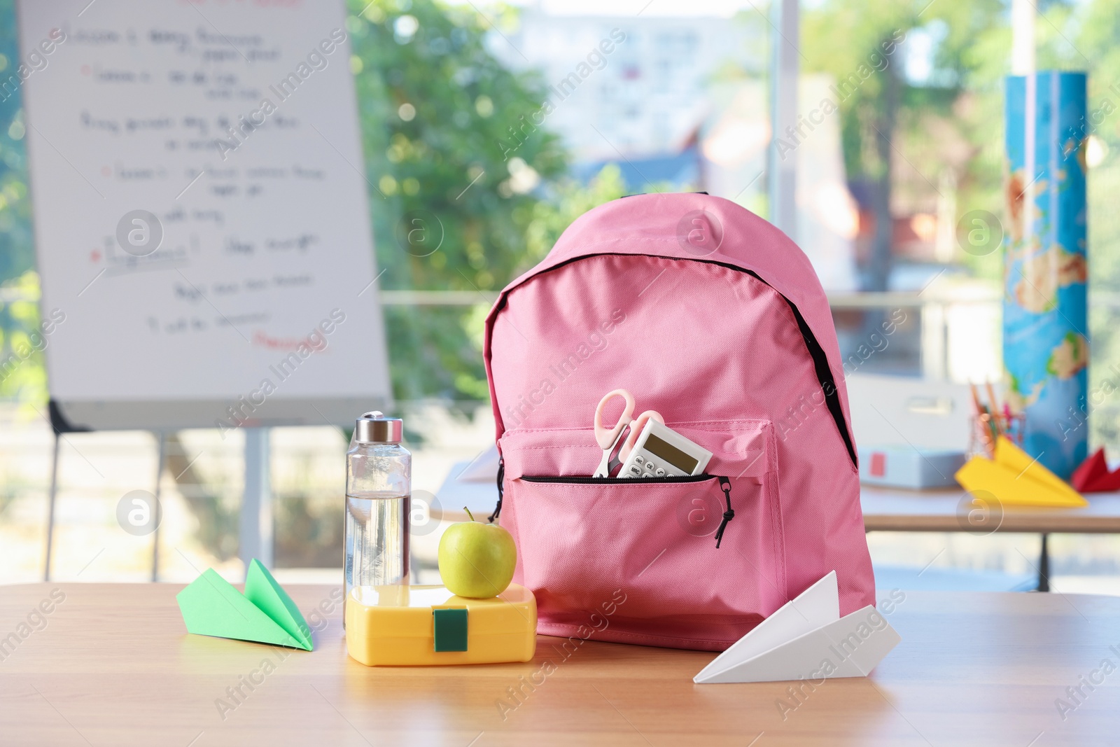Backpack with different stationery, lunchbox, apple, bottle of water and paper planes on desk in classroom Photo of Backpack with different stationery, lunchbox, apple, bottle of water and paper planes on desk in classroom