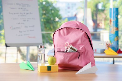 Photo of Backpack with different stationery, lunchbox, apple, bottle of water and paper planes on desk in classroom