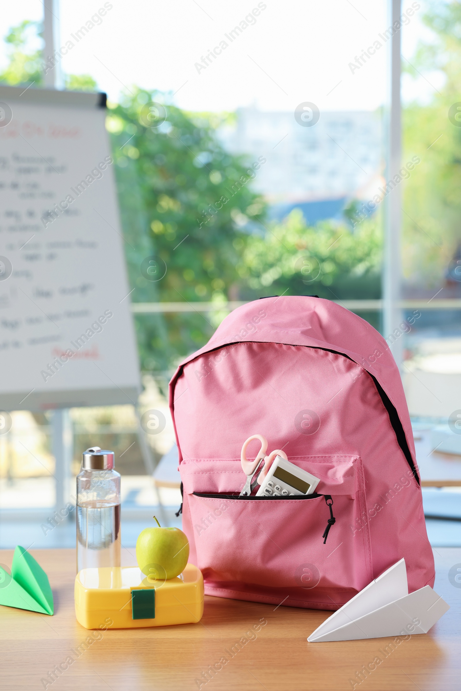 Photo of Backpack with different stationery, lunchbox, apple, bottle of water and paper planes on desk in classroom