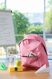 Photo of Backpack with different stationery, lunchbox, apple, bottle of water and paper planes on desk in classroom