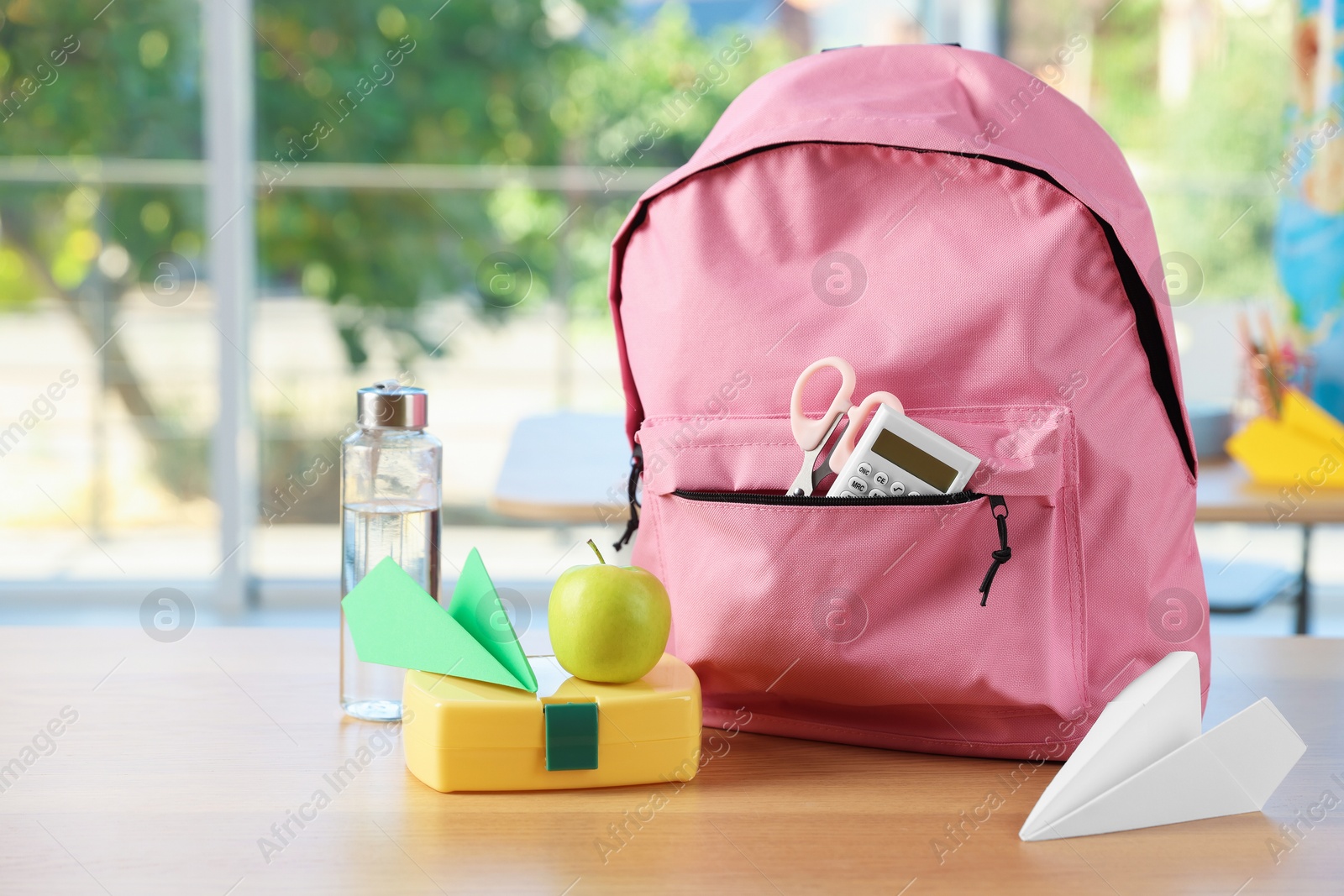 Photo of Backpack with different stationery, lunchbox, apple, bottle of water and paper planes on desk in classroom