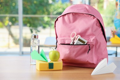 Photo of Backpack with different stationery, lunchbox, apple, bottle of water and paper planes on desk in classroom