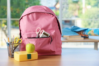 Photo of Backpack with different stationery, lunchbox and apple on desk in classroom