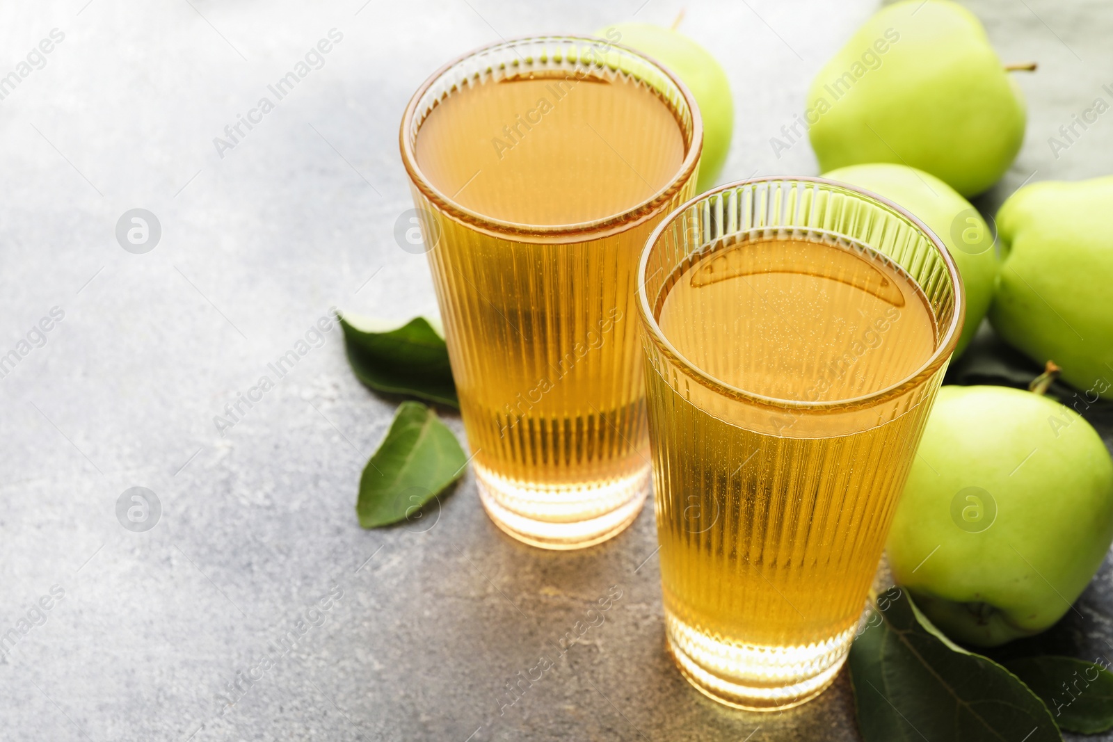 Photo of Delicious cider in glasses, apples and leaves on grey textured table, closeup. Space for text