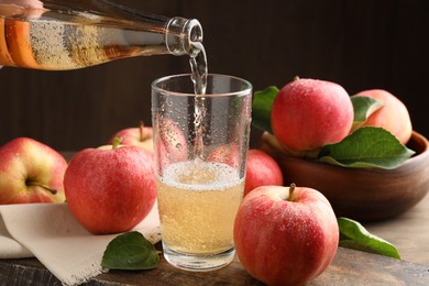 Photo of Pouring delicious apple cider from bottle into glass at wooden table against dark background, closeup