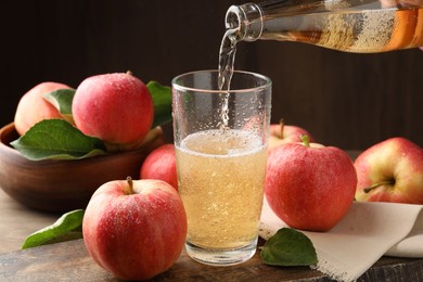 Photo of Pouring delicious apple cider from bottle into glass at wooden table against dark background, closeup
