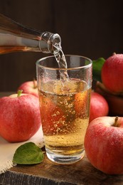 Photo of Pouring delicious apple cider from bottle into glass at wooden table against dark background, closeup