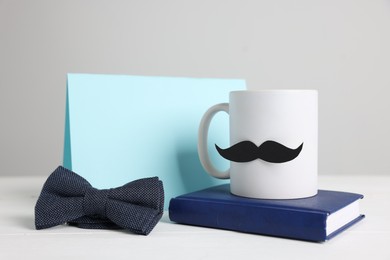 Photo of Happy Father's Day. Cup with paper moustache, bowtie, book and card on white wooden table