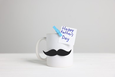 Photo of Happy Father's Day. Cup with paper moustache and card on white wooden table