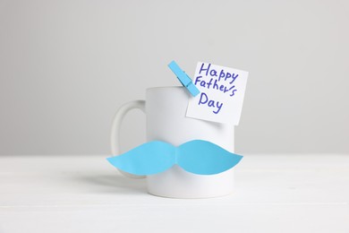 Photo of Happy Father's Day. Cup with paper moustache and card on white wooden table, closeup