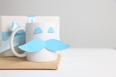 Photo of Happy Father's Day. Cup with paper face and gift box on white wooden table, closeup. Space for text
