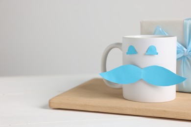 Photo of Happy Father's Day. Cup with paper face and gift box on white wooden table, closeup. Space for text