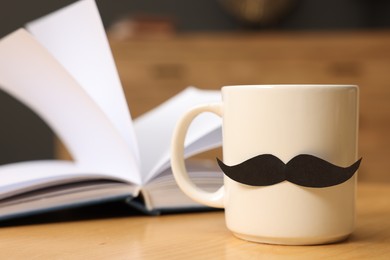 Photo of Happy Father's Day. Cup with paper moustache and book on wooden table indoors, closeup