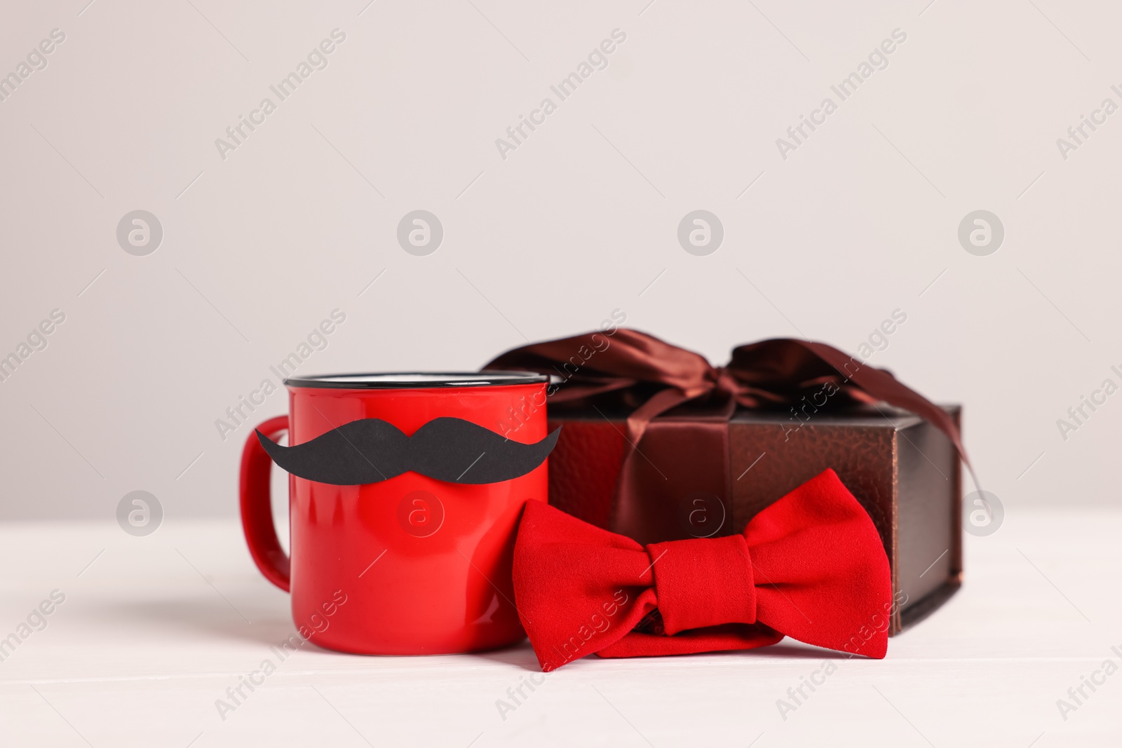 Happy Father's Day. Cup with paper moustache, bowtie and gift box on white wooden table, closeup Photo of Happy Father's Day. Cup with paper moustache, bowtie and gift box on white wooden table, closeup