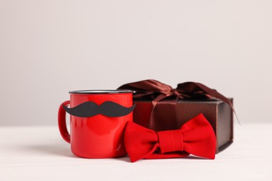 Photo of Happy Father's Day. Cup with paper moustache, bowtie and gift box on white wooden table, closeup