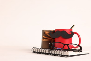 Photo of Happy Father's Day. Cup with paper moustache, notebook, glasses and gift box on white wooden table, space for text