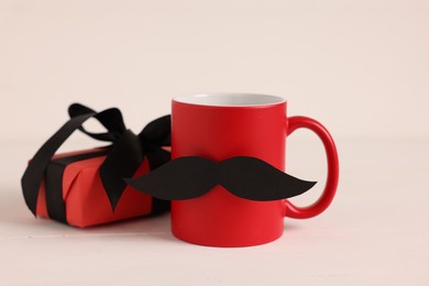 Photo of Happy Father's Day. Cup with paper moustache and gift box on white wooden table, closeup