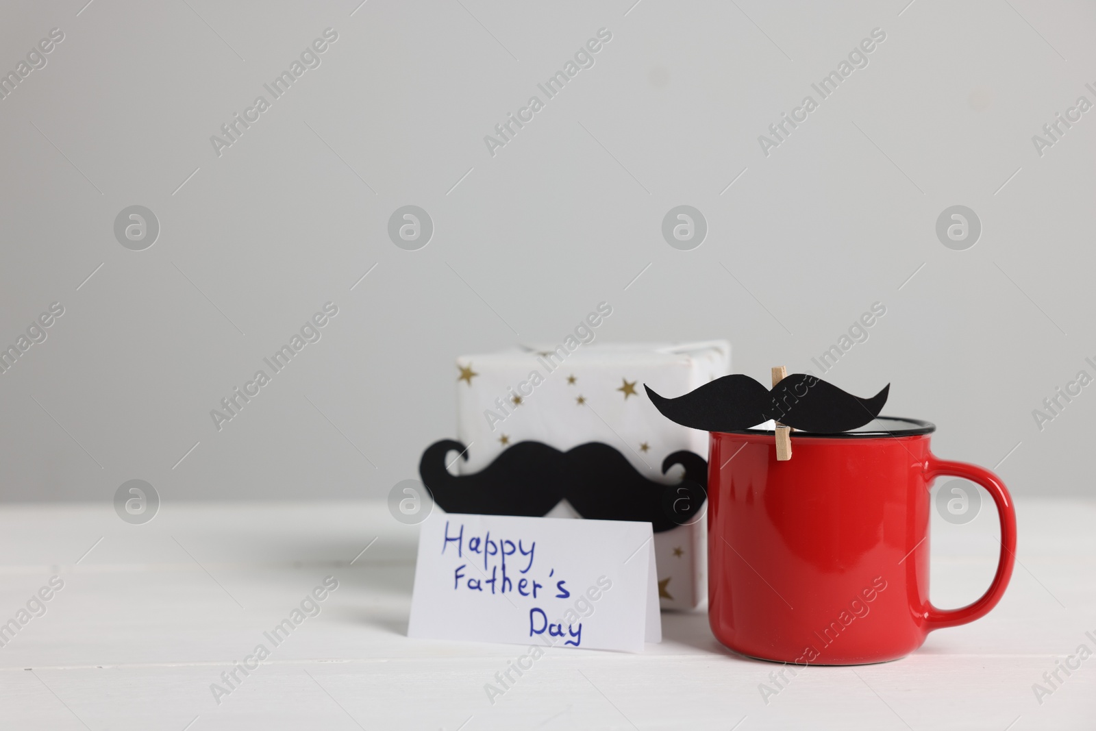 Happy Father's Day. Cup with paper moustaches, card and gift box on white wooden table, space for text Photo of Happy Father's Day. Cup with paper moustaches, card and gift box on white wooden table, space for text