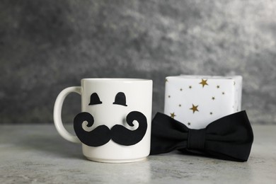 Photo of Happy Father's Day. Cup with paper face, gift box and bowtie on light grey table, closeup