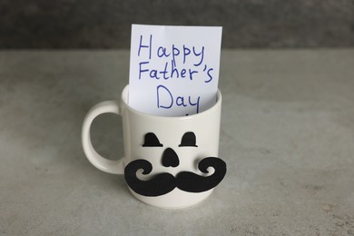 Photo of Happy Father's Day. Cup with paper face and card on light grey table, closeup