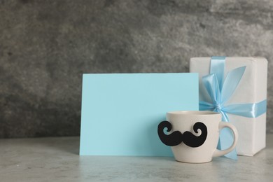 Photo of Happy Father's Day. Cup with paper moustache, gift box and card on light grey table, space for text