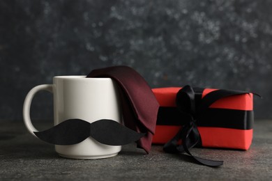 Photo of Happy Father's Day. Cup with paper moustache, gift box and tie on black table, closeup