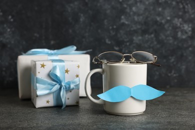 Photo of Happy Father's Day. Cup with paper moustache, gift boxes and glasses on black table, closeup