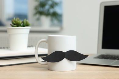 Photo of Happy Father's Day. Cup with paper moustache and laptop on wooden table, closeup