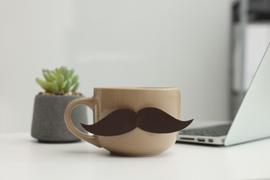 Photo of Happy Father's Day. Cup with paper moustache and laptop on white table indoors, closeup