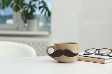 Photo of Happy Father's Day. Cup with paper moustache, books and glasses on white table indoors, space for text