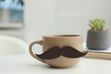 Photo of Happy Father's Day. Cup with paper moustache and books on white table indoors, closeup