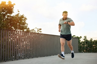 Photo of Athletic young man in sportswear running and checking fitness tracker outdoors, space for text
