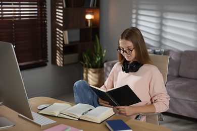 Photo of Teenage girl reading book while studying online at table indoors