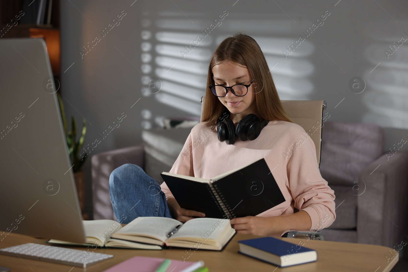 Teenage girl reading book while studying online at table indoors Photo of Teenage girl reading book while studying online at table indoors