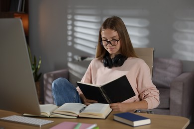 Photo of Teenage girl reading book while studying online at table indoors