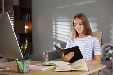 Photo of Teenage girl reading book while studying online at table indoors