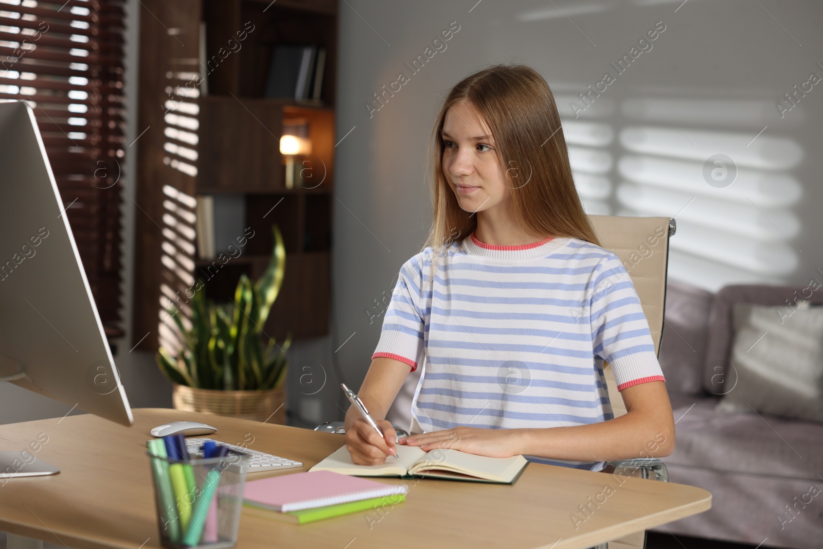 Teenage girl taking notes while studying online at table indoors Photo of Teenage girl taking notes while studying online at table indoors