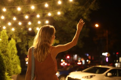 Photo of Young woman catching taxi on city street at night, back view