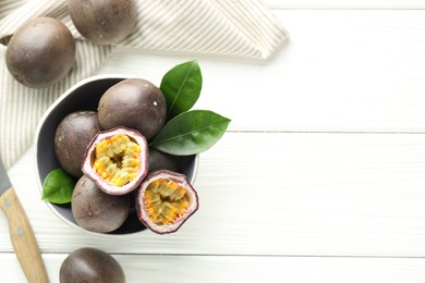 Photo of Fresh passion fruits in bowl, green leaves and knife on white wooden table, flat lay. Space for text