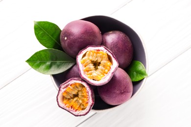Photo of Fresh passion fruits and green leaves in bowl on white wooden table, top view