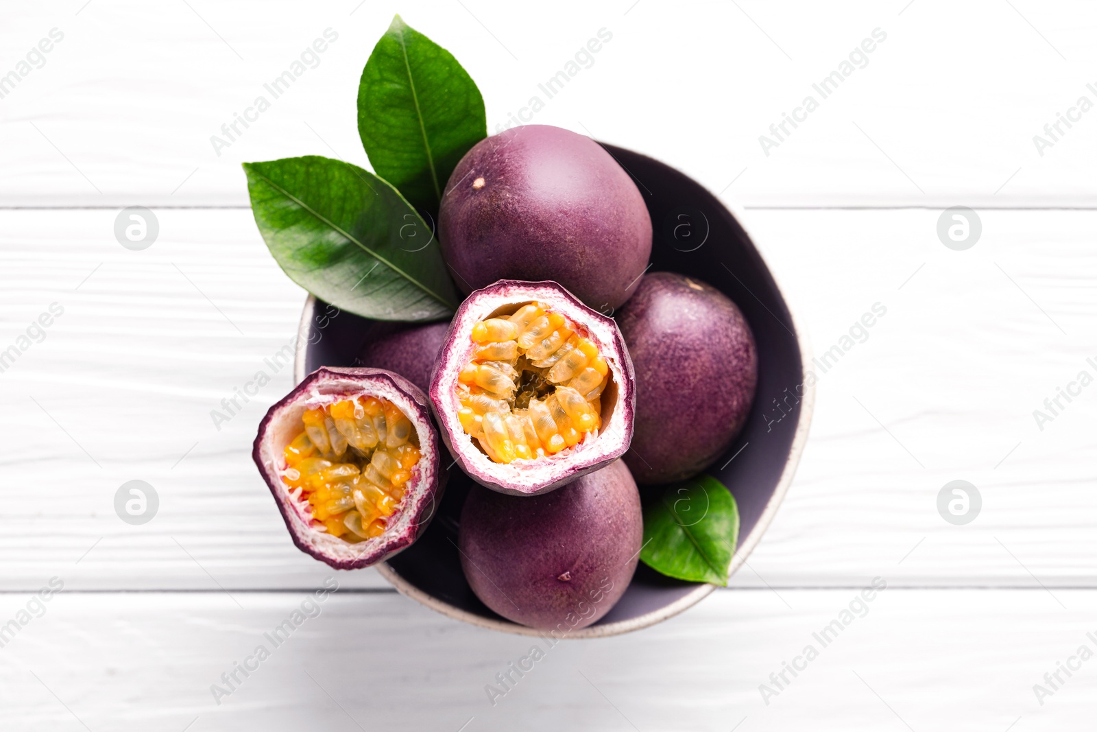 Photo of Fresh passion fruits and green leaves in bowl on white wooden table, top view