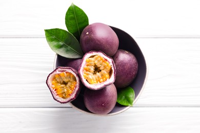 Photo of Fresh passion fruits and green leaves in bowl on white wooden table, top view
